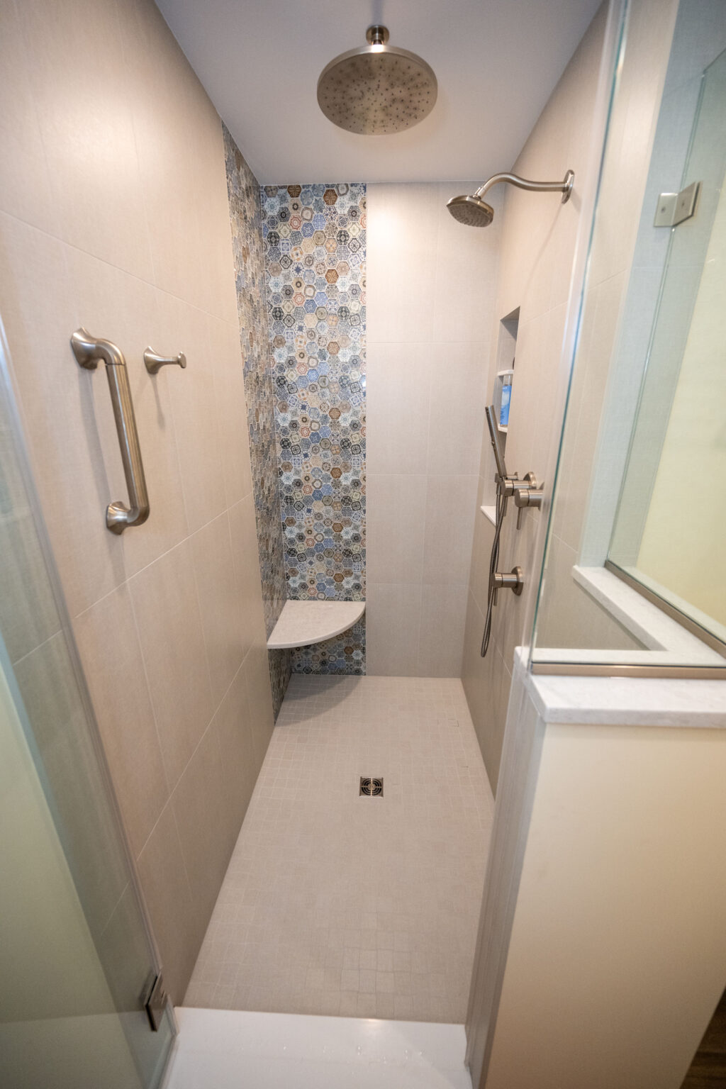 A modern shower with beige tiles, a decorative mosaic strip, a rainfall showerhead, wall-mounted handheld shower, a small corner bench, and a glass door.