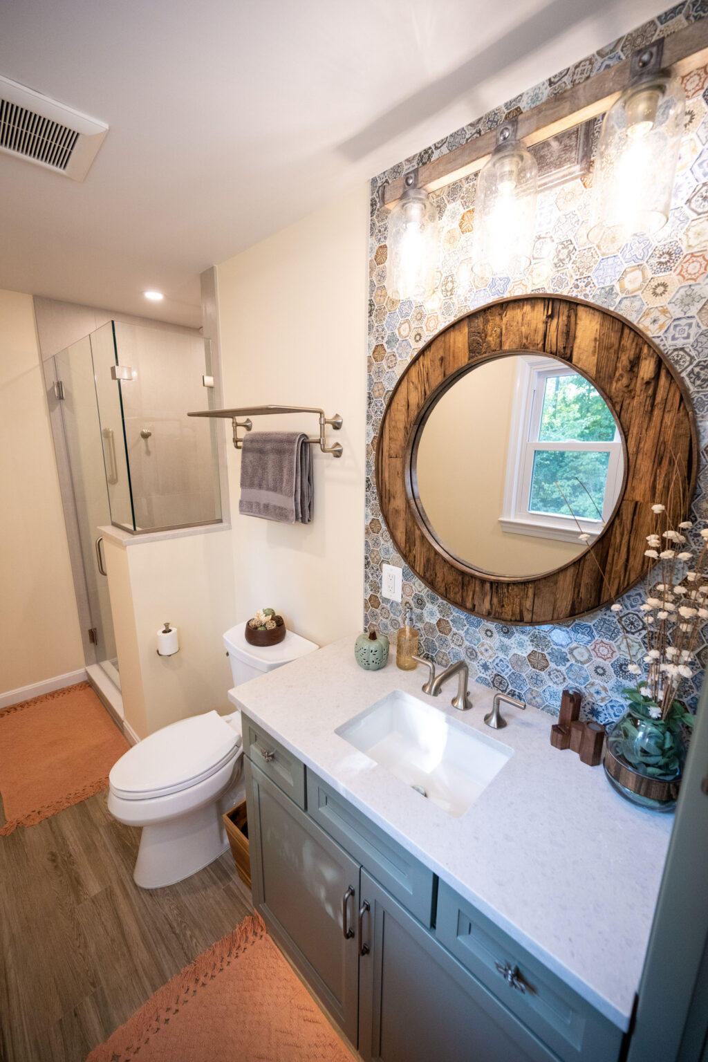 Bathroom with a round mirror above a vanity, decorative tile backsplash, glass shower enclosure, and wooden flooring.