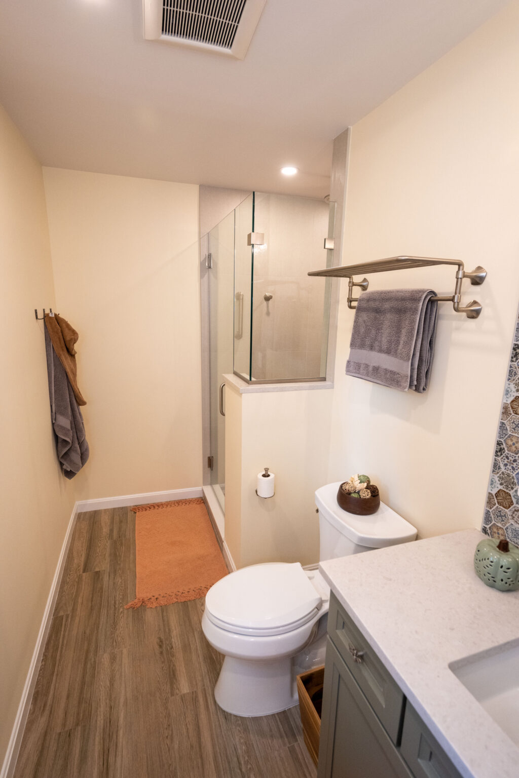 Bathroom with a glass-enclosed shower, toilet, and vanity. Towels hang on racks, and a small mat lies on the wooden floor. Walls are light-colored, and there's a decorative tile section.