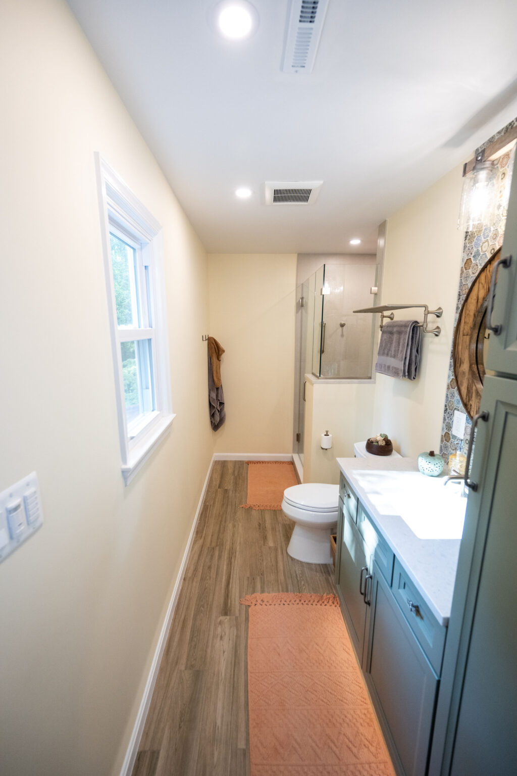 Narrow bathroom with wood-look flooring, white walls, a window, toilet, shower, and sink with marble countertop. Orange bathmats are on the floor.