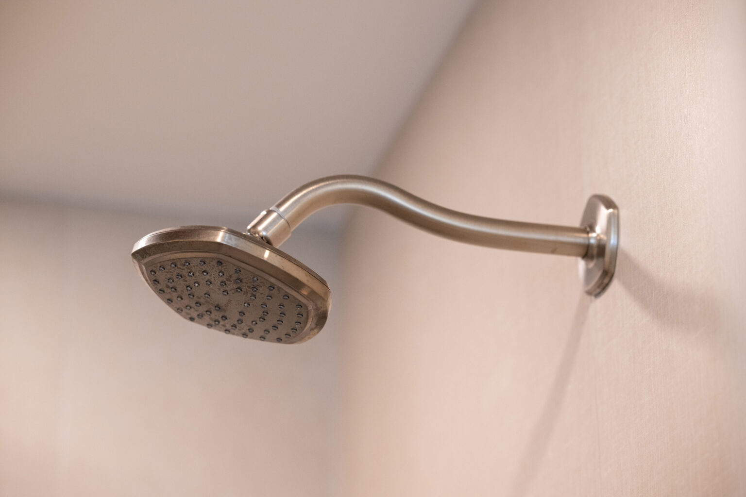 A modern metallic showerhead attached to a curved arm is mounted on a beige wall.