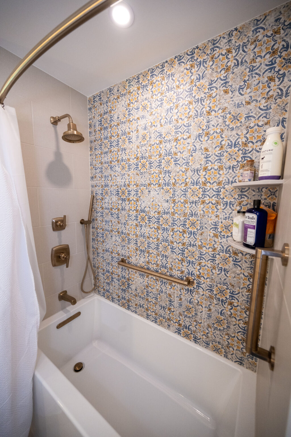 Bathroom with a white bathtub, brass fixtures, and a showerhead. The wall features colorful mosaic tiles. Bath products are on a shelf.