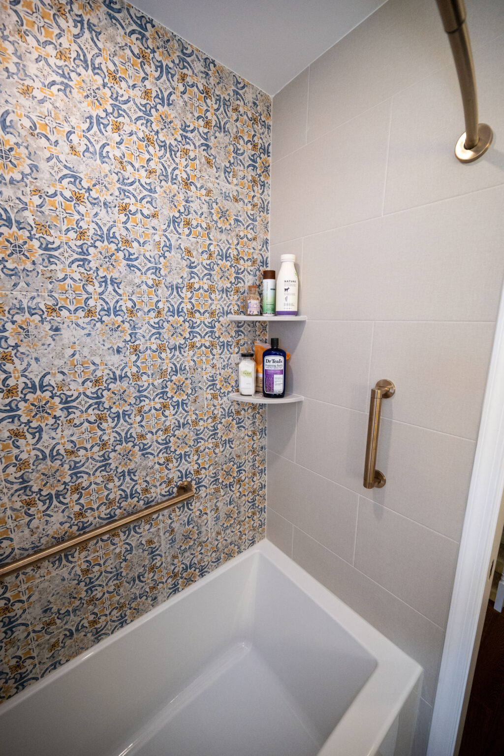 Bathroom with a bathtub, ornate blue and yellow tiled wall, white shelves holding toiletries, and a curved shower rod with a bronze bar for support.