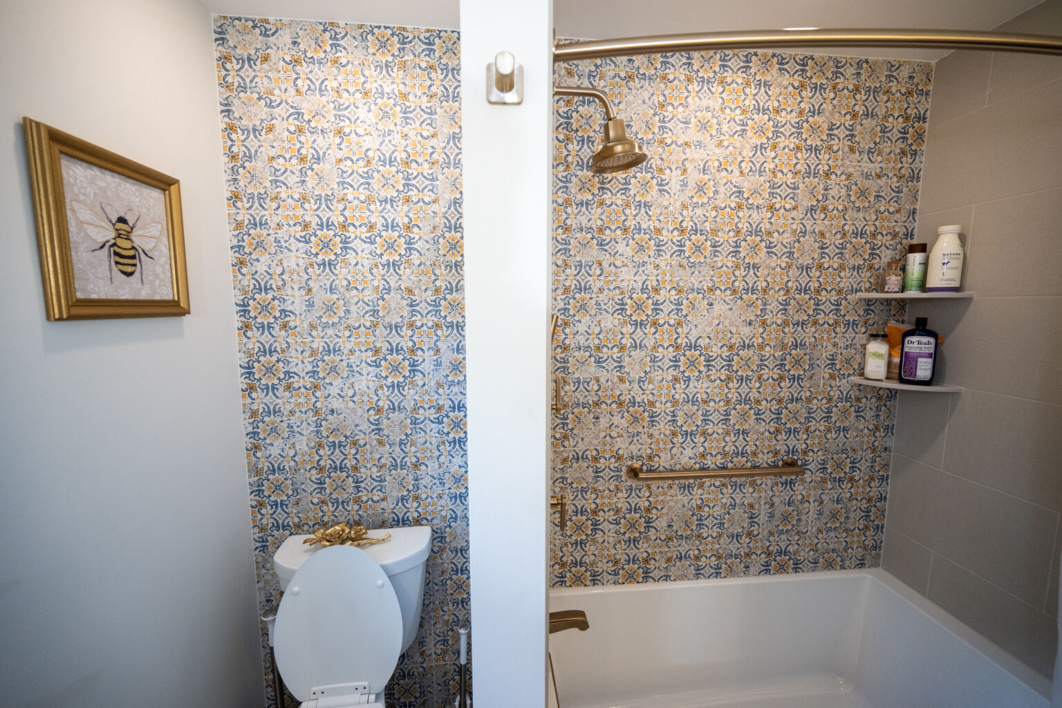 Bathroom with a tiled shower, gold fixtures, and a bathtub. There's a toilet with an open lid, and a bee-themed picture on the wall. Shelves hold toiletries.