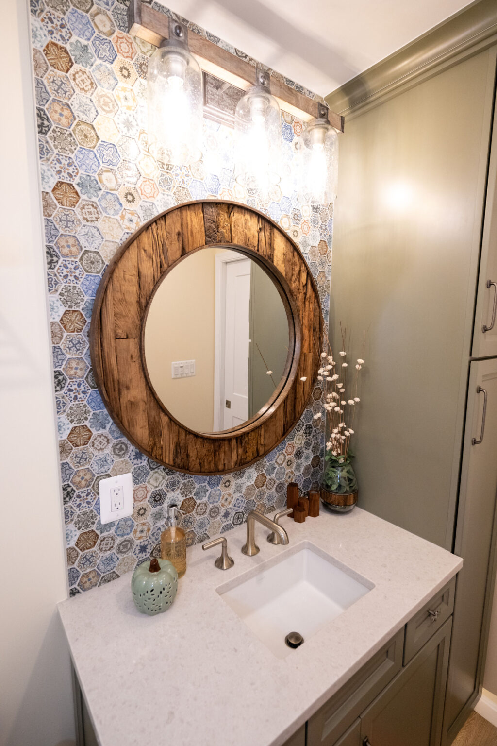 A bathroom with a round wooden mirror, patterned tile backsplash, sink with dual faucets, and decorative items on the counter.