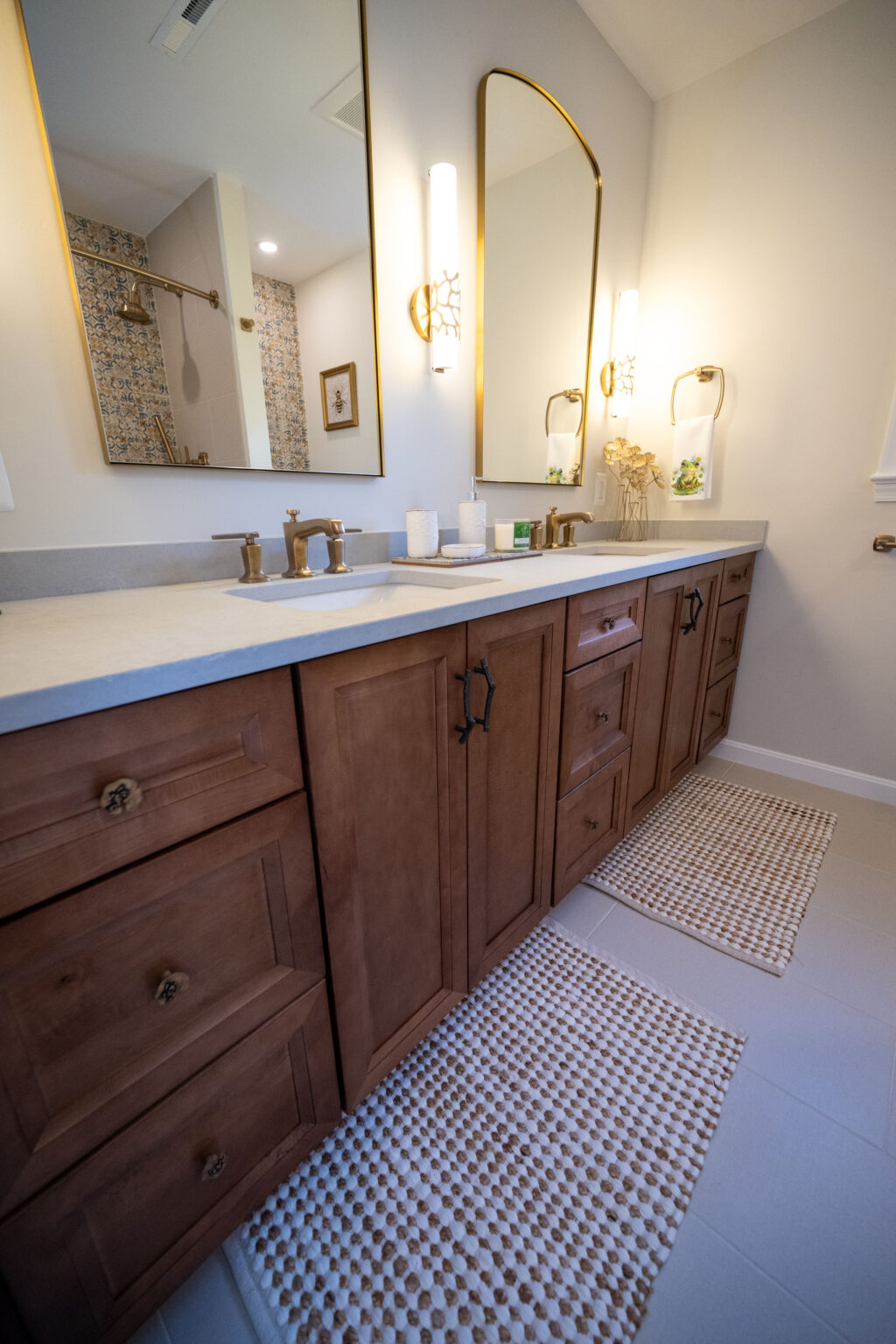 A bathroom with a double sink vanity, wooden cabinets, two large mirrors, wall sconces, two bath mats, and a view of a shower with a glass door.