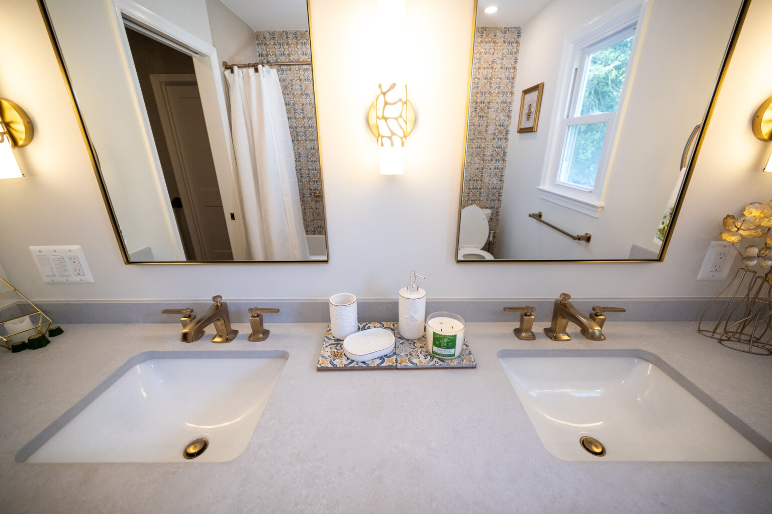 A bathroom features a double sink vanity with brass faucets, large mirrors, a light fixture between mirrors, and decorative items. A shower curtain and a window are visible in the background.