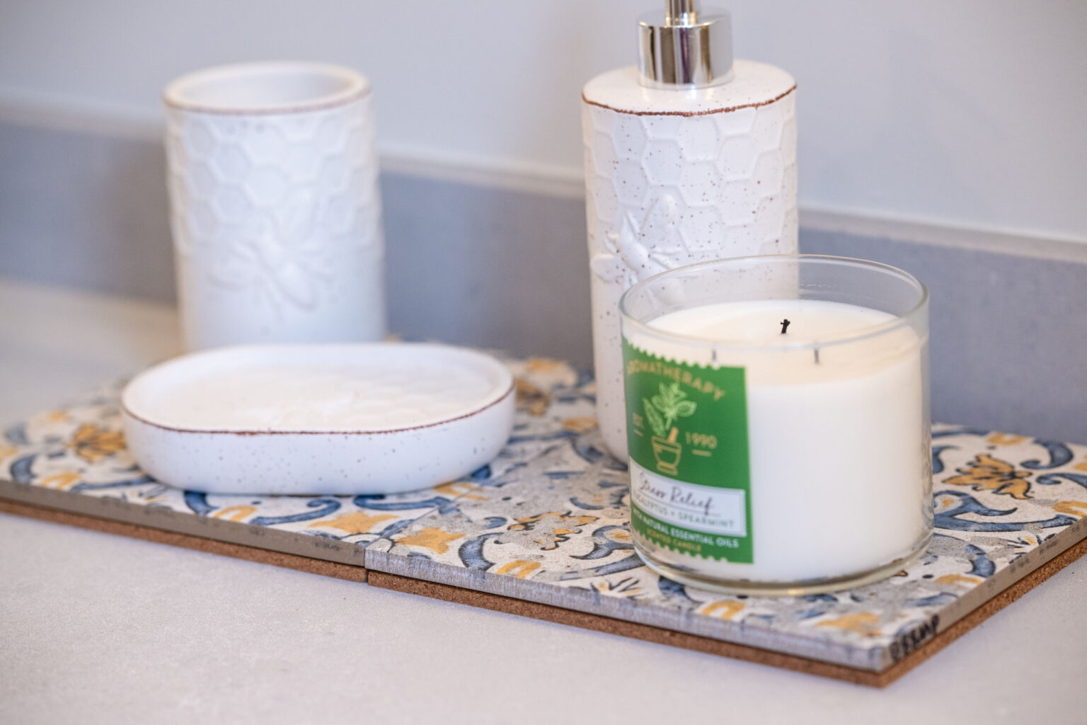 Ceramic bathroom set with a white candle, soap dish, and dispenser on a patterned tray.
