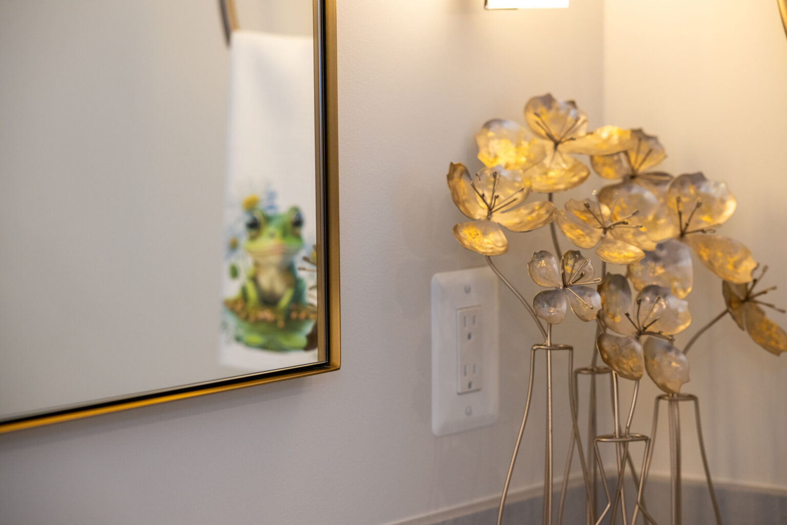 Decorative flowers and a light switch on a wall beside a mirror reflecting a frog illustration on a towel.