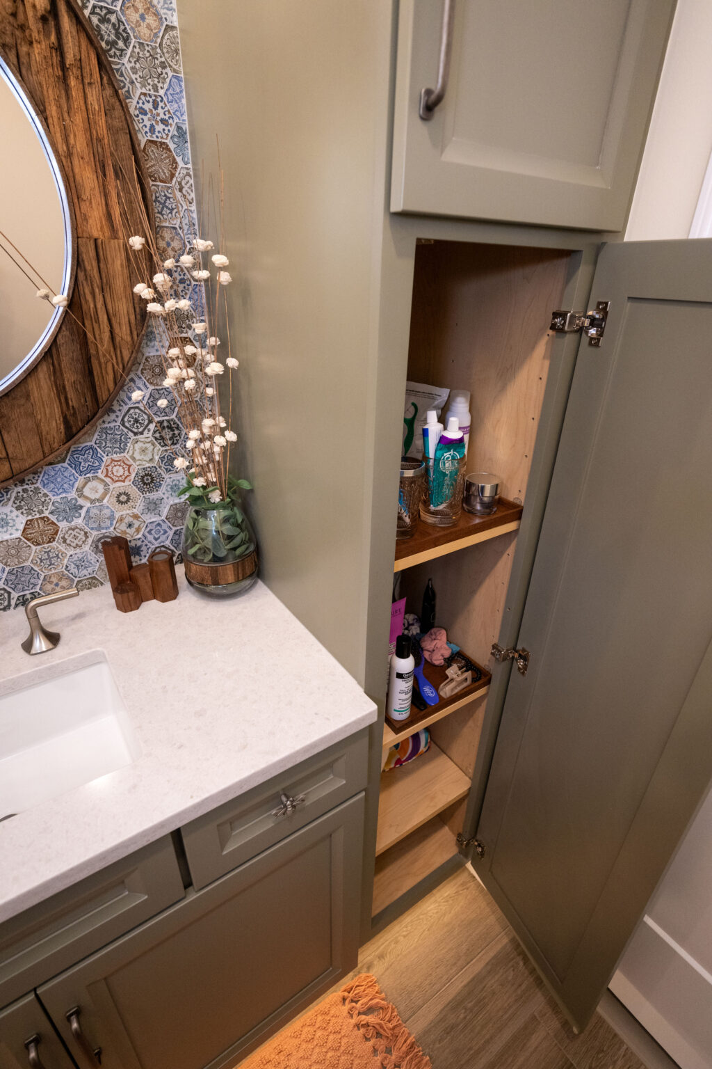 Bathroom cabinet open revealing shelves with toiletries and cleaning supplies next to a sink with a mirror and decorative vase.