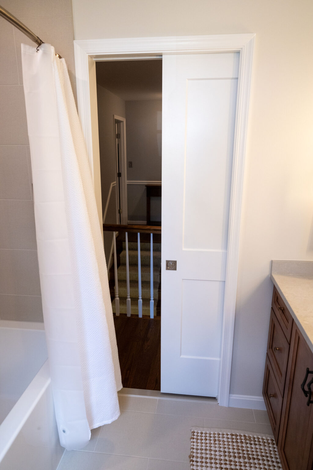 Bathroom with white sliding door open, revealing a staircase. Bathtub with a white curtain on the left and a wooden vanity with a countertop on the right.