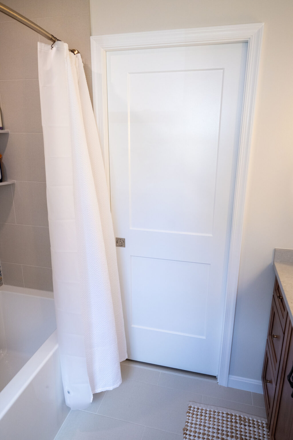 White bathroom with a closed white door, white bathtub, and shower curtain. Beige tiled floor and a corner of a wooden vanity with countertop visible.