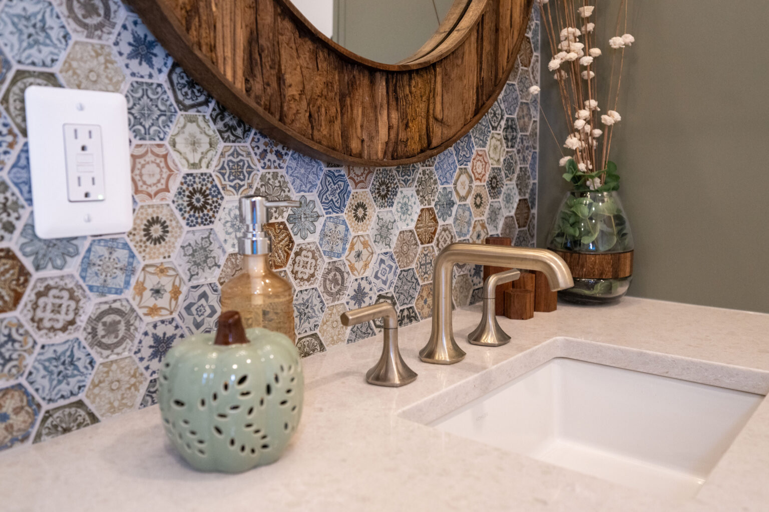 Bathroom sink area with a hexagonal patterned backsplash, round wooden mirror, silver faucet, green apple-shaped decoration, soap dispenser, and vase with dried flowers.