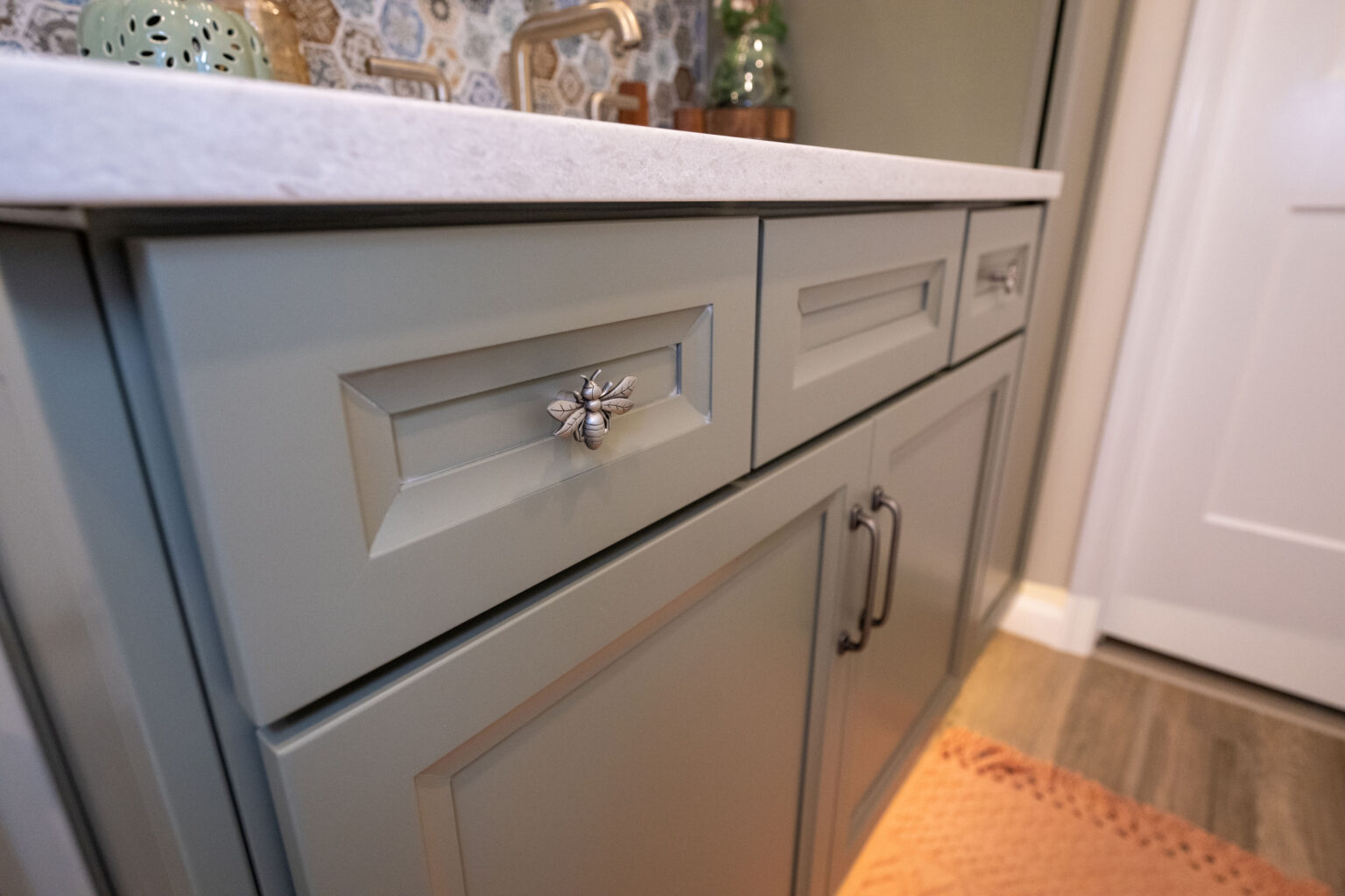 A bathroom vanity with light green cabinets, silver handles, and a white countertop is shown. Decor includes a potted plant and a patterned wall behind.