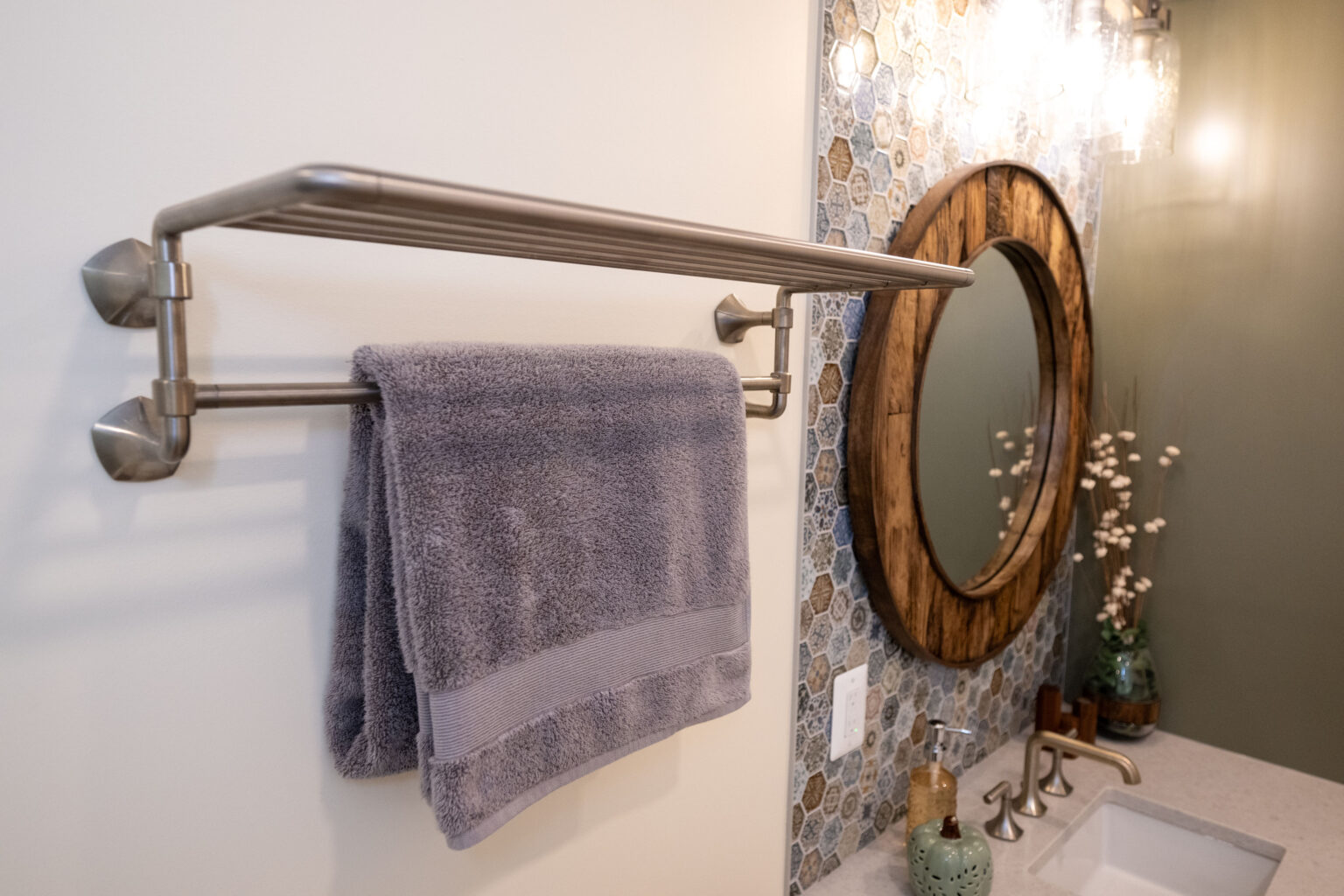 A bathroom features a gray towel hanging on a double towel bar, a round mirror with a wooden frame, mosaic tiled wall, light fixture, and part of a sink with a faucet and decorative plant.