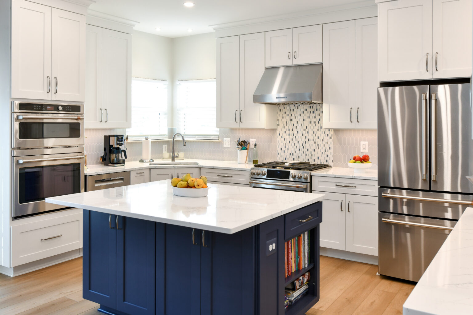 105-Shakley-DSC_2076-3 Modern kitchen with white cabinets, stainless steel appliances, a blue island with a white countertop, and a fruit bowl. Light wood flooring and geometric backsplash add contrast.
