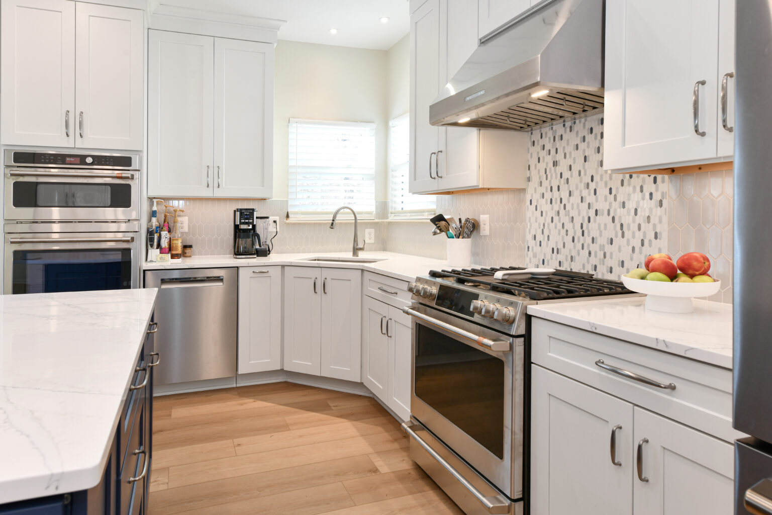 106-Shakley-DSC_2172 Modern kitchen with white cabinets, stainless steel appliances, marble countertops, and wooden floor. A fruit bowl is on the counter, and a window with blinds is above the sink.