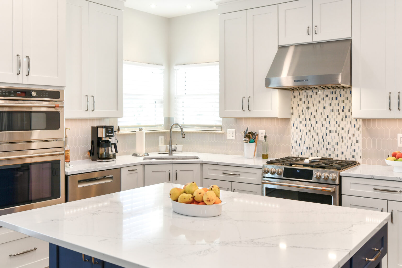 107-Shakley-DSC_2084-2 Modern kitchen with white cabinets, stainless steel appliances, and a marble island with a fruit bowl. Mosaic backsplash and windows provide natural light.