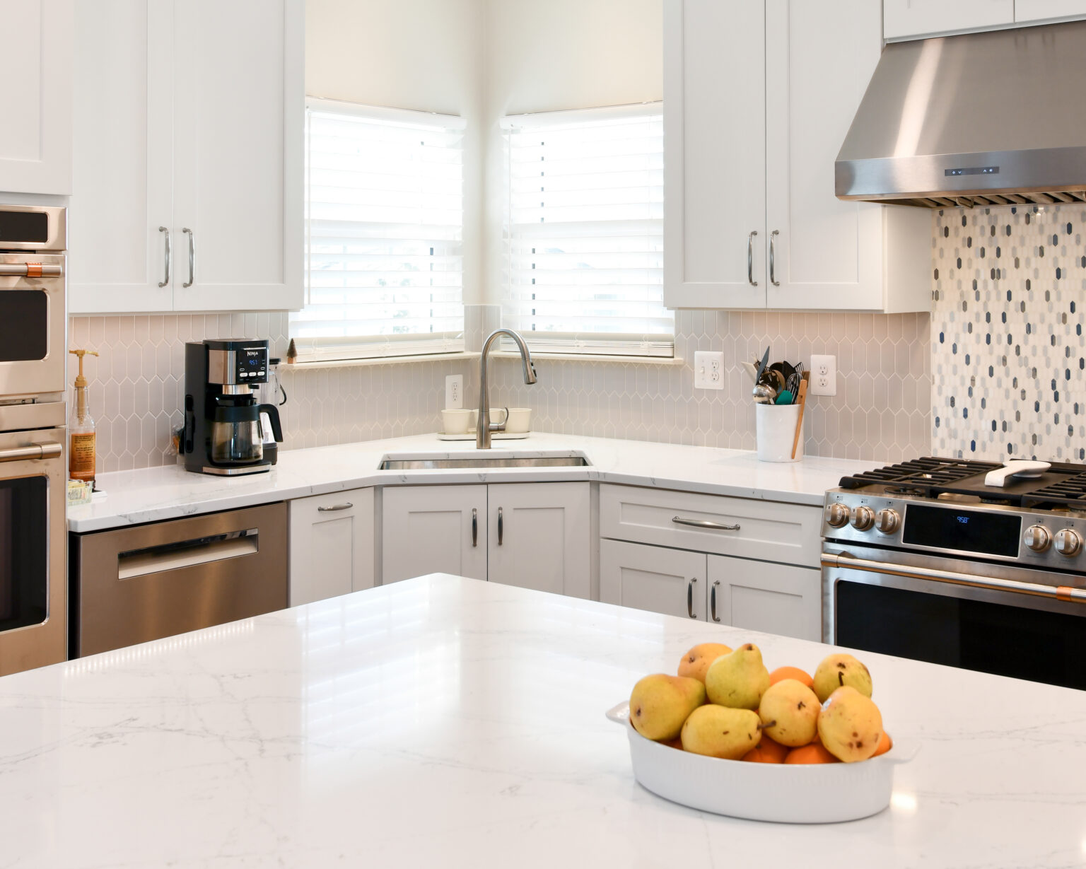 108-Shakley-DSC_2095 Modern kitchen with stainless steel appliances, white cabinets, and a marble countertop. A bowl of apples and pears sits on the central island. Bright natural light from window blinds.