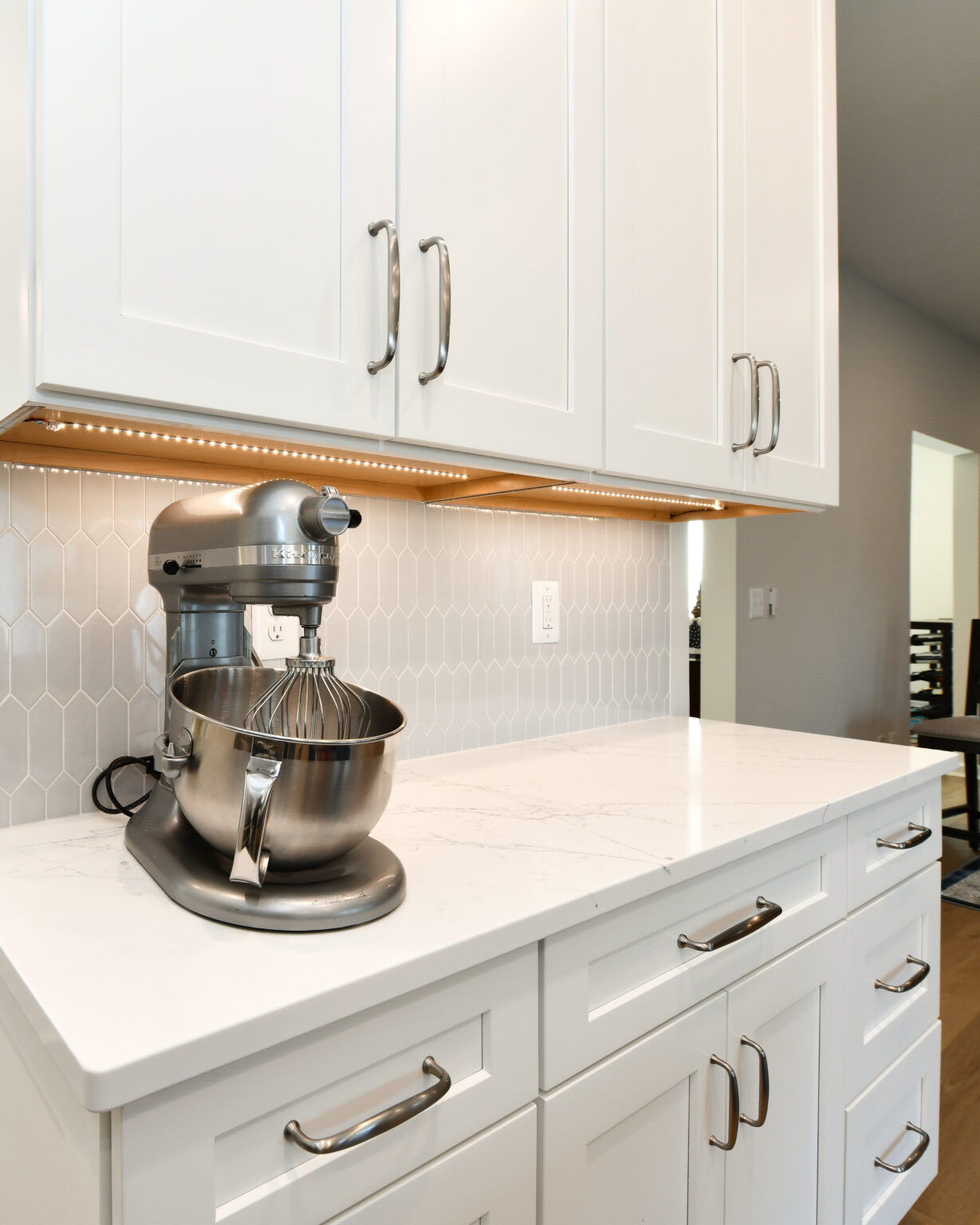 109-Shakley-DSC_2134 A silver stand mixer on a white countertop beneath white cabinets with metal handles, featuring a hexagonal tile backsplash and under-cabinet lighting.