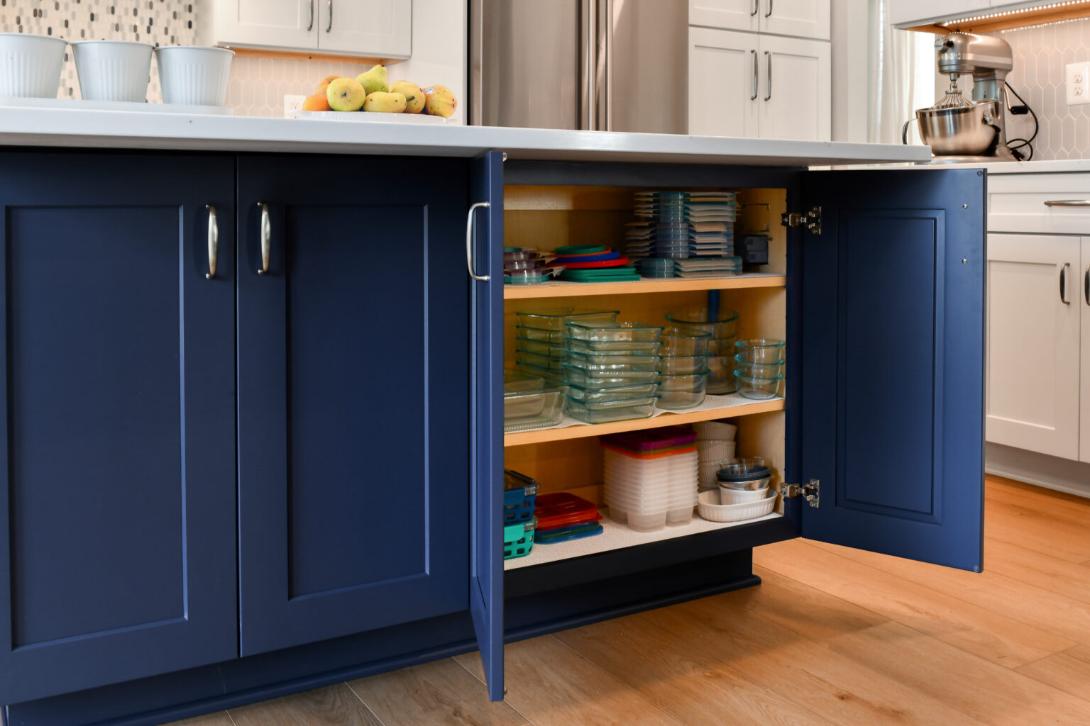 110-Shakley-DSC_2237 Open kitchen cabinet with blue doors displaying organized glass and plastic containers on wooden shelves, under a countertop with a bowl of apples.