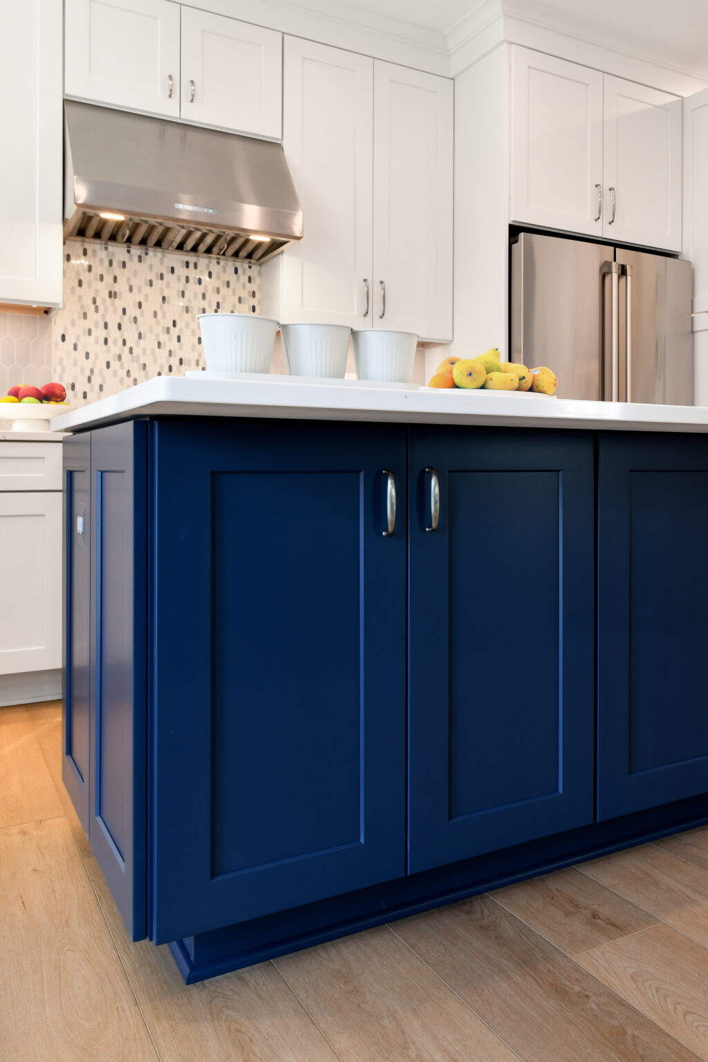 111-Shakley-DSC_2241 Modern kitchen with white cabinets, stainless steel appliances, a blue island, and a wooden floor. The countertop holds three white containers and a bunch of bananas.