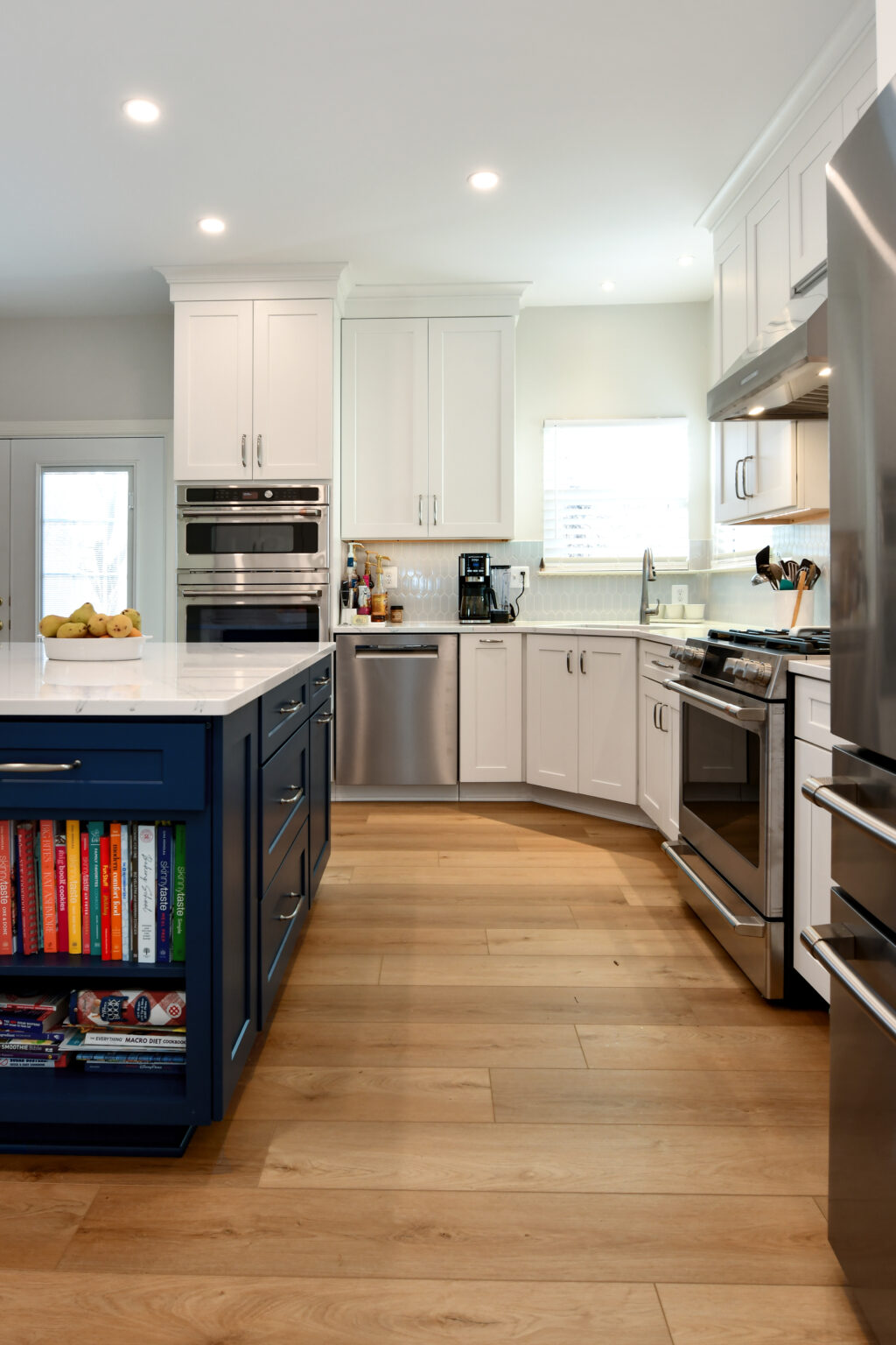 112-Shakley-DSC_2150 Modern kitchen with white cabinets, stainless steel appliances, a blue island featuring bookshelves, and wooden flooring.