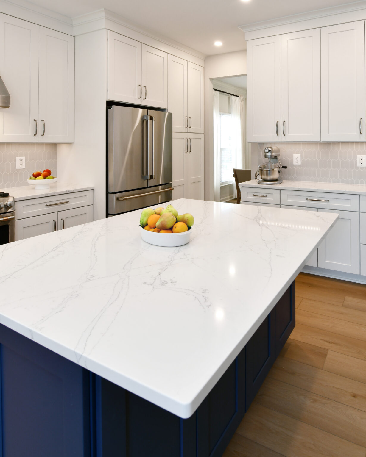 113-Shakley-DSC_2120 Modern kitchen with white cabinets, stainless steel appliances, and a large island with a white marble countertop. A fruit bowl sits at the center of the island.