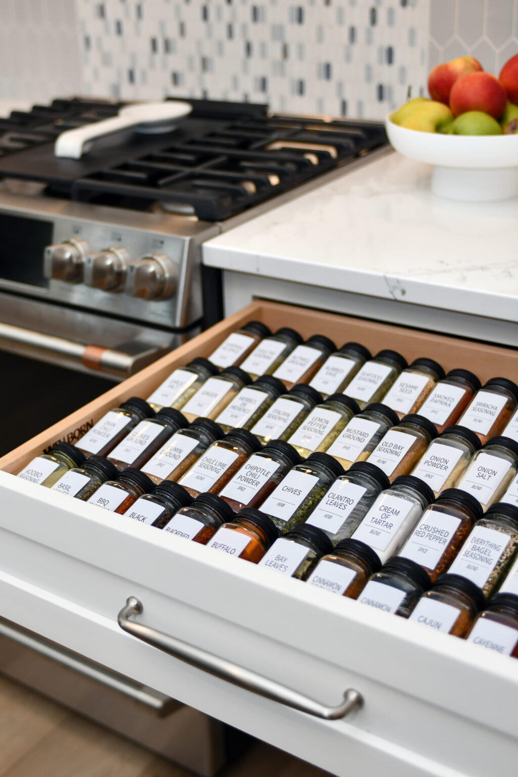 115-Shakley-DSC_2177 Open kitchen drawer with neatly organized spice jars labeled with names, next to a stove and a bowl of fruit on the counter.
