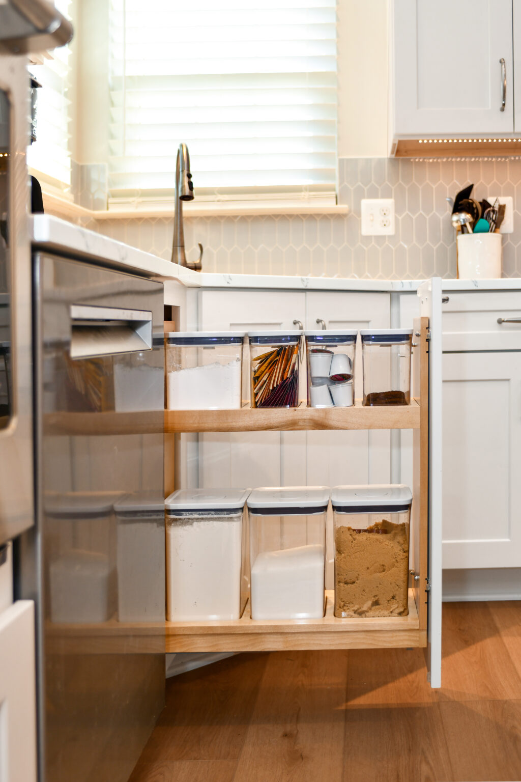 118-Shakley-DSC_2195 A kitchen cabinet with a pull-out shelf holding various containers filled with different ingredients, placed beside a dishwasher and a sink under a window.