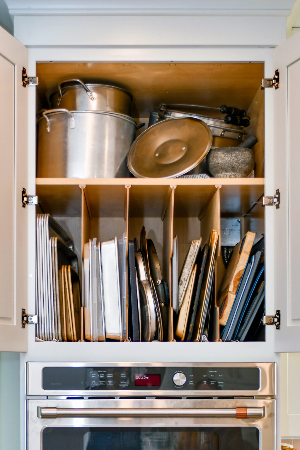 119-Shakley-DSC_2205-3 A kitchen cabinet above an oven is open, showing neatly organized baking trays, pans, and cooking pots stored on shelves.