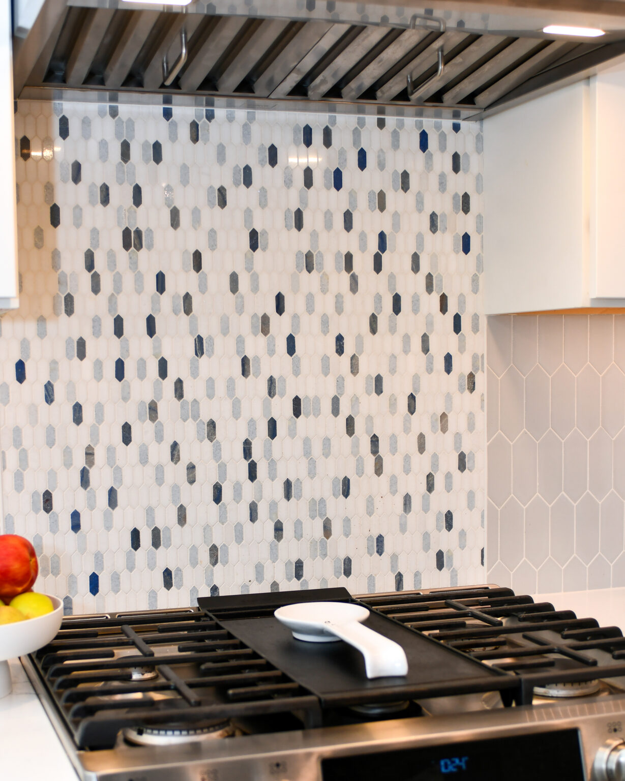 120-Shakley-DSC_2207 Kitchen stovetop with a black griddle and a white teapot, set against a backsplash of small, multicolored hexagonal tiles. A fruit bowl with apples is on the counter to the left.
