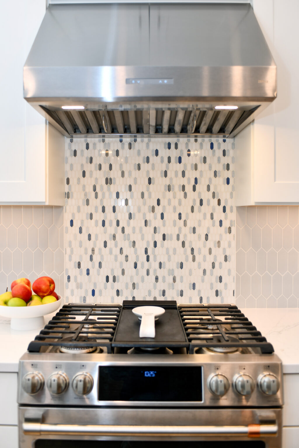 121-Shakley-DSC_2214 Modern kitchen with stainless steel stove and hood, decorative mosaic tile backsplash, and a bowl of fruit on the counter.