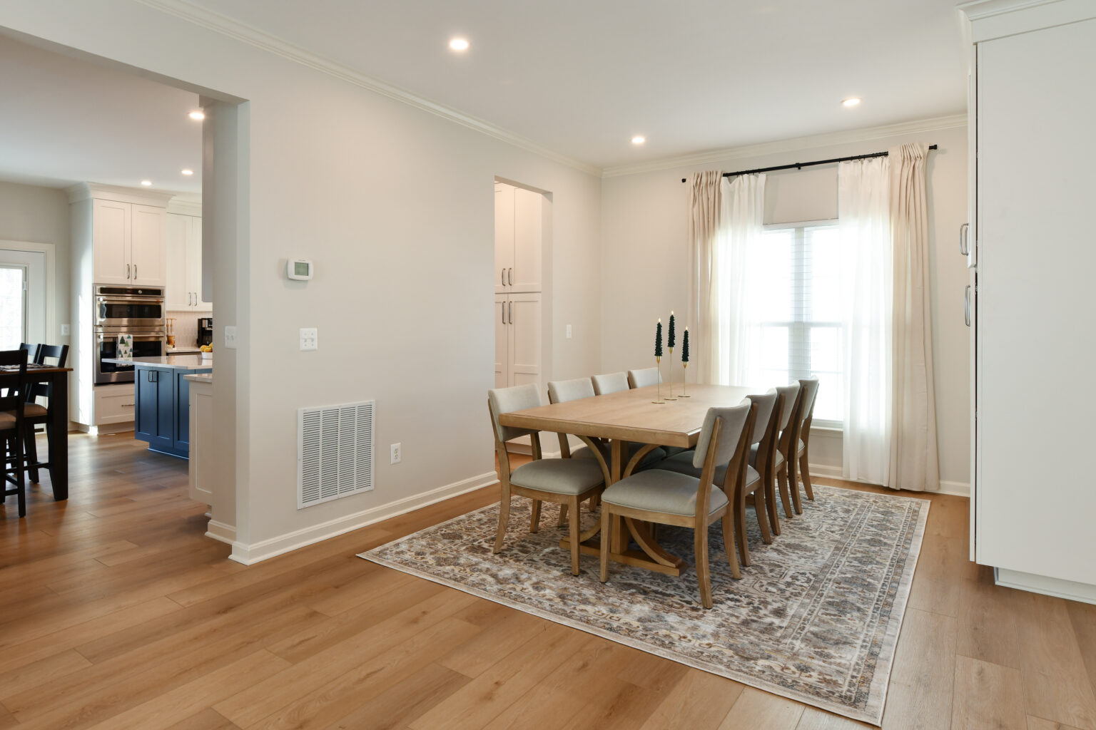 123-Shakley-DSC_2285 Dining room with a wooden table and eight chairs on a patterned rug. Light walls, ceiling lights, and a window with curtains. Kitchen area visible in the background.