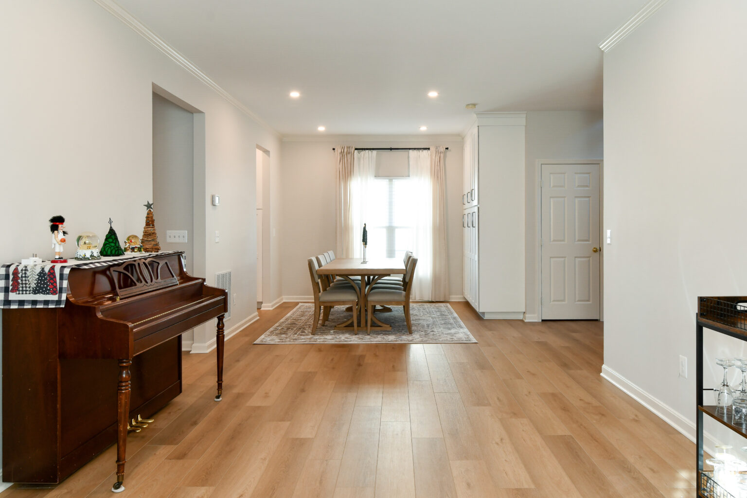 124-Shakley-DSC_2288 Dining room with wooden floor, a table and chairs on a rug, a piano on the left, and white walls with a window under white curtains.