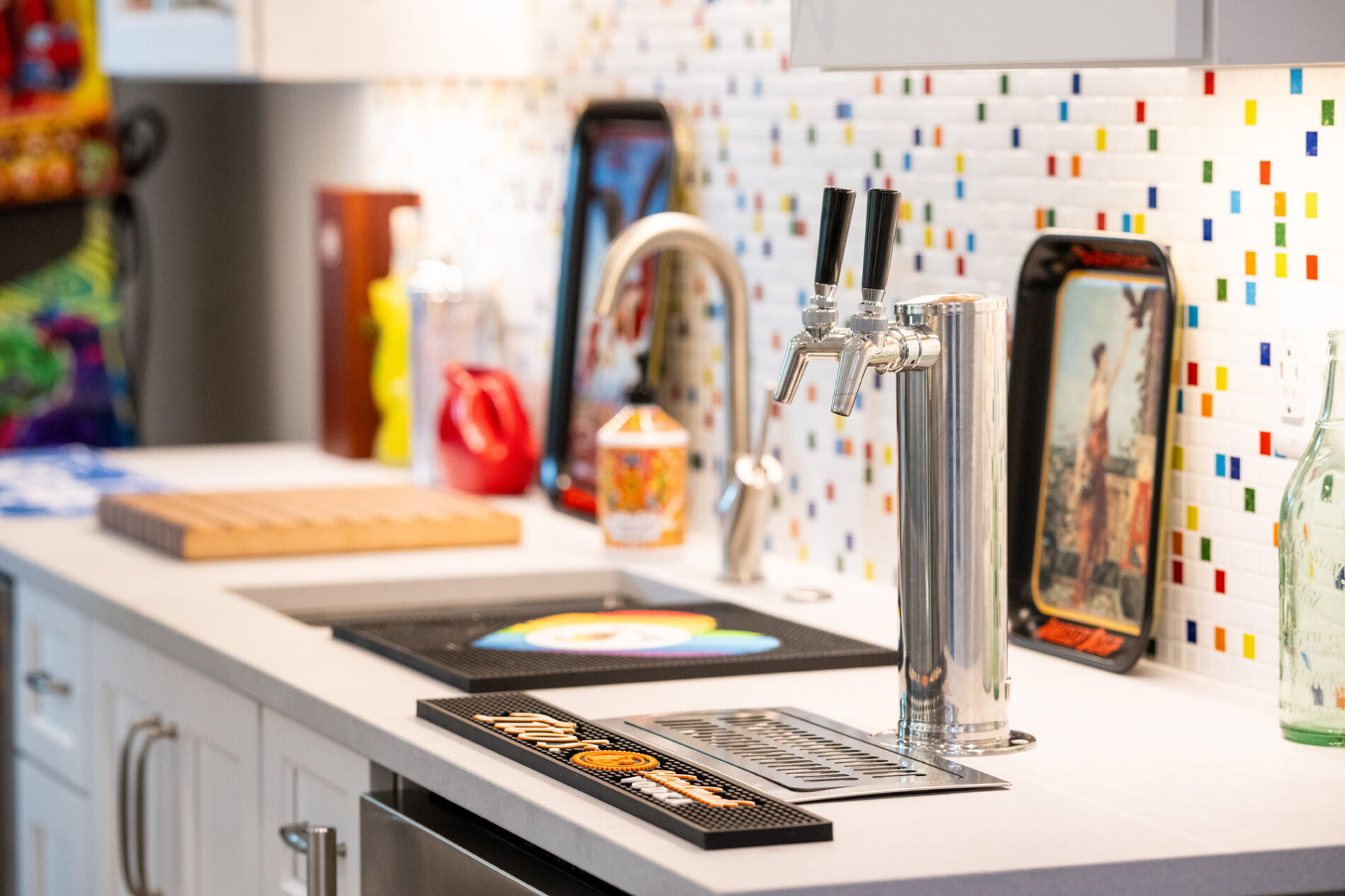 Modern kitchen counter with a dual beer tap, colorful mosaic backsplash, trays, cutting board, sink, and various decorative items.