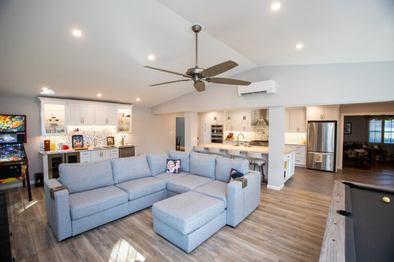 A modern open-concept living space with a gray sectional sofa, pool table, bar area, and kitchen, featuring wood flooring and recessed lighting.