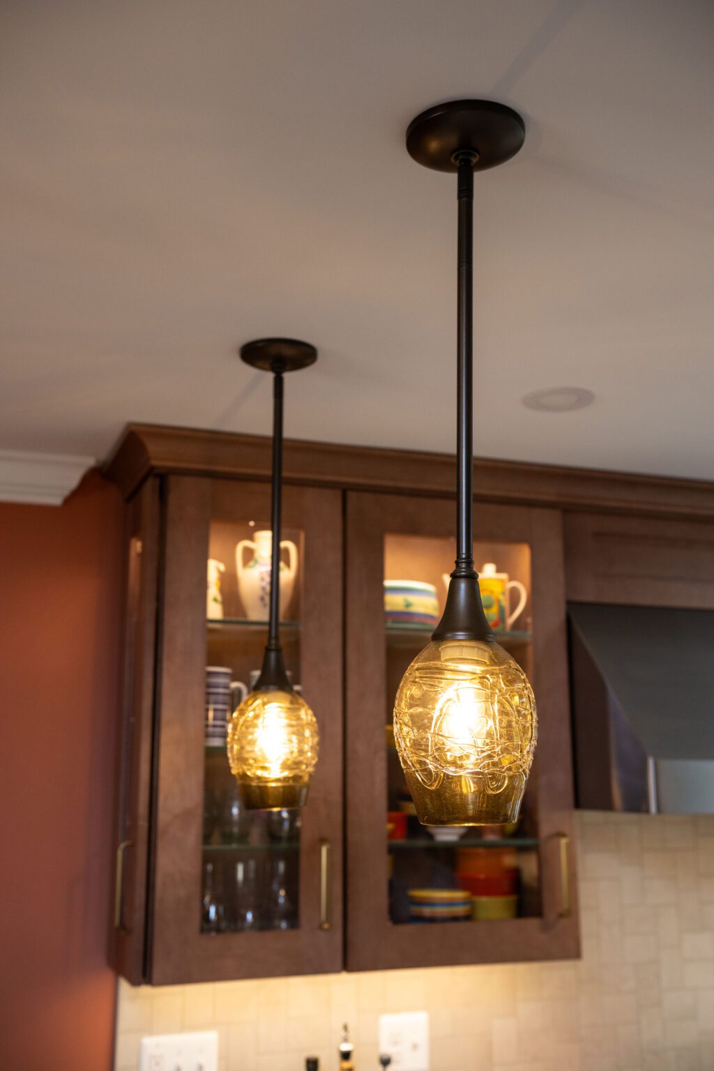 Image by Lesnick Photo Two glass pendant lights hang from the ceiling in a kitchen, with wooden cabinets and shelves displaying mugs and dishes in the background.