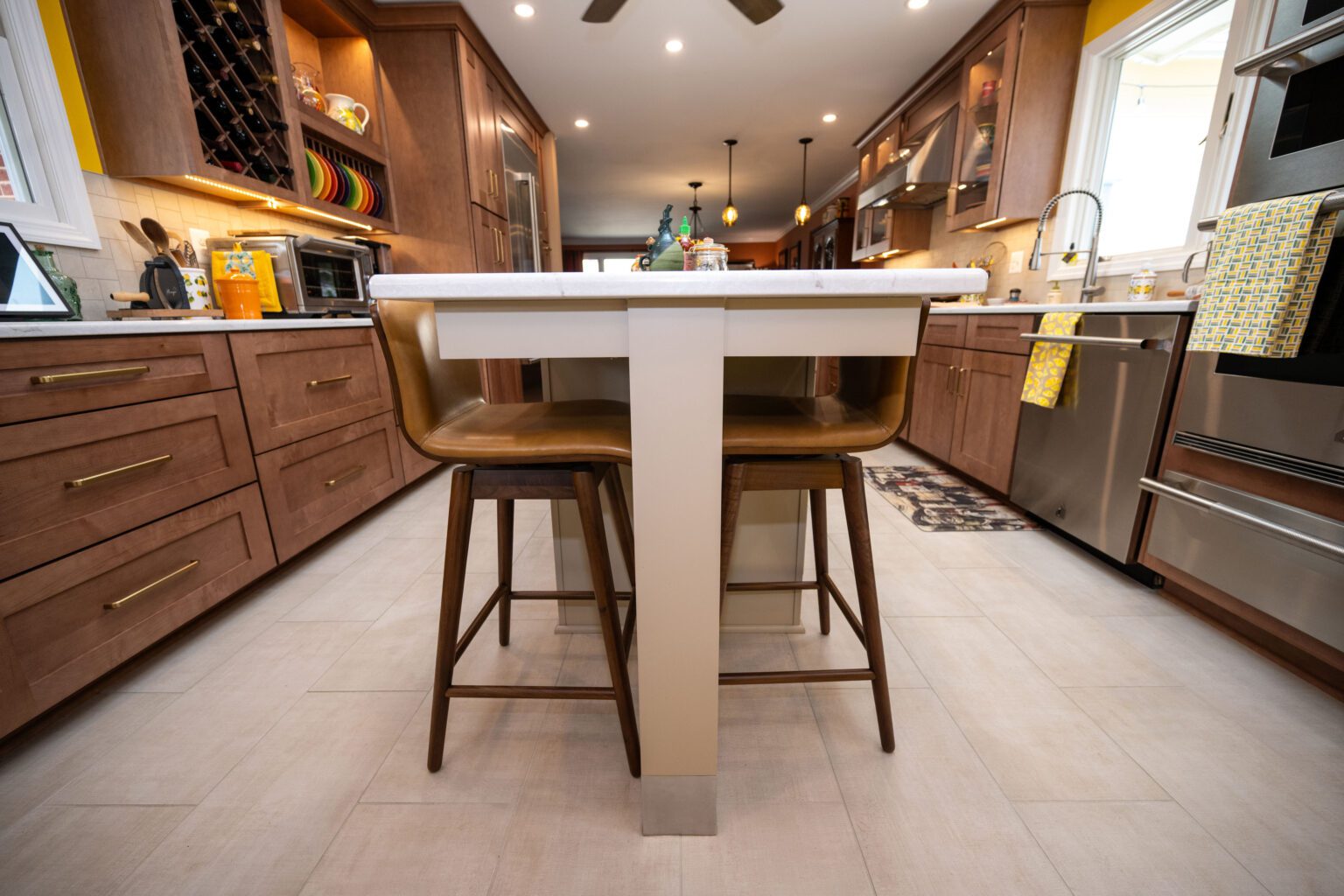 Image by Lesnick Photo Modern kitchen with wooden cabinets, stainless steel appliances, a central island with two brown chairs, and various kitchen items on countertops and shelves.