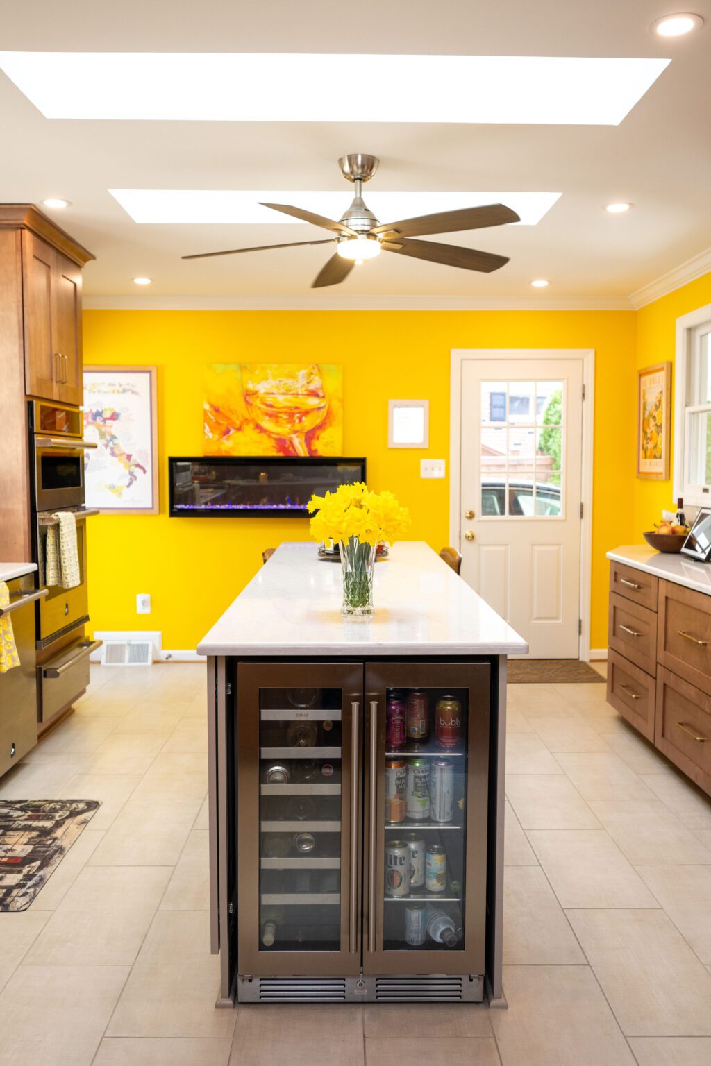 Image by Lesnick Photo Modern kitchen with bright yellow walls, a ceiling fan, skylights, artwork, and an island featuring a beverage fridge and a vase of yellow flowers.