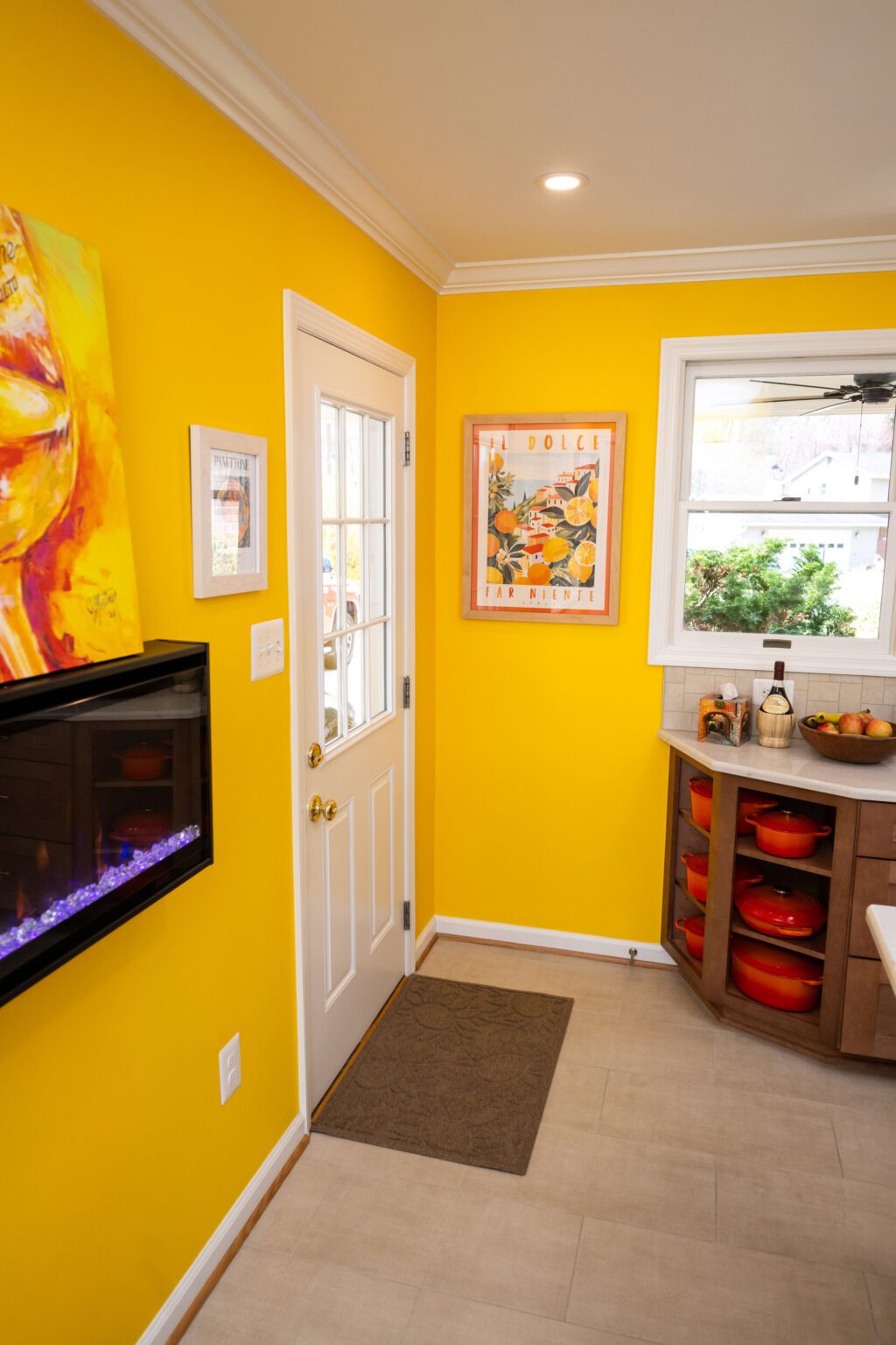Image by Lesnick Photo A brightly lit kitchen with yellow walls, a white door, colorful artwork, a countertop with bowls and bottles, and a small electric fireplace.