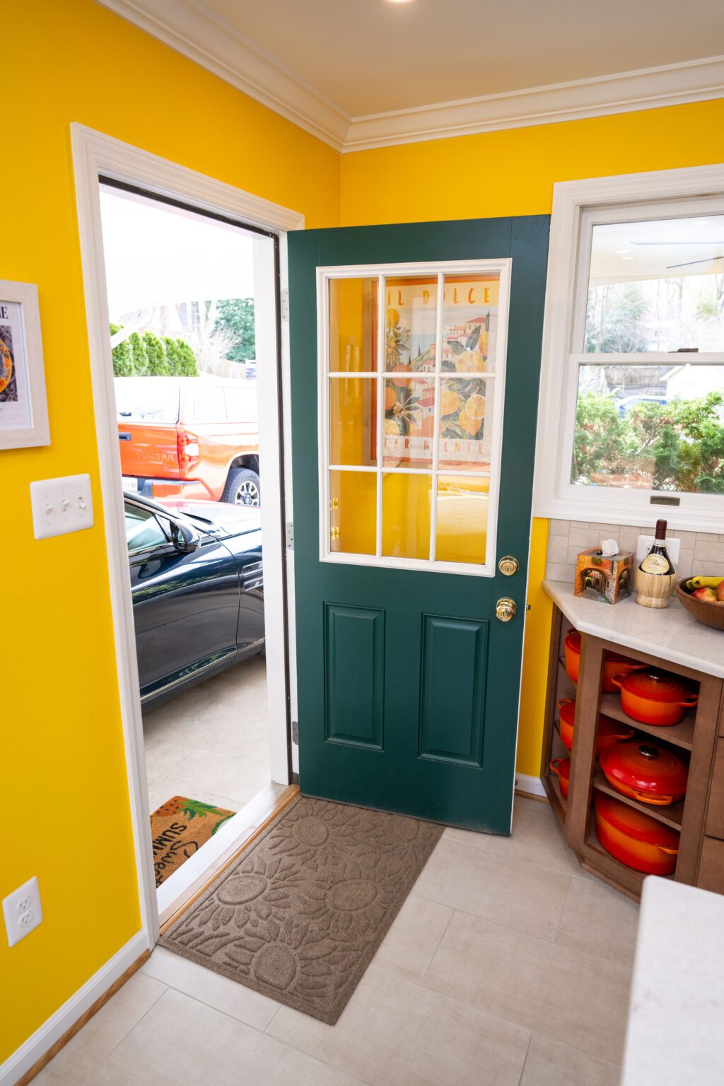 Image by Lesnick Photo A green door with glass panels stands open to a driveway with two parked cars; the kitchen has bright yellow walls and a cabinet with red bowls.