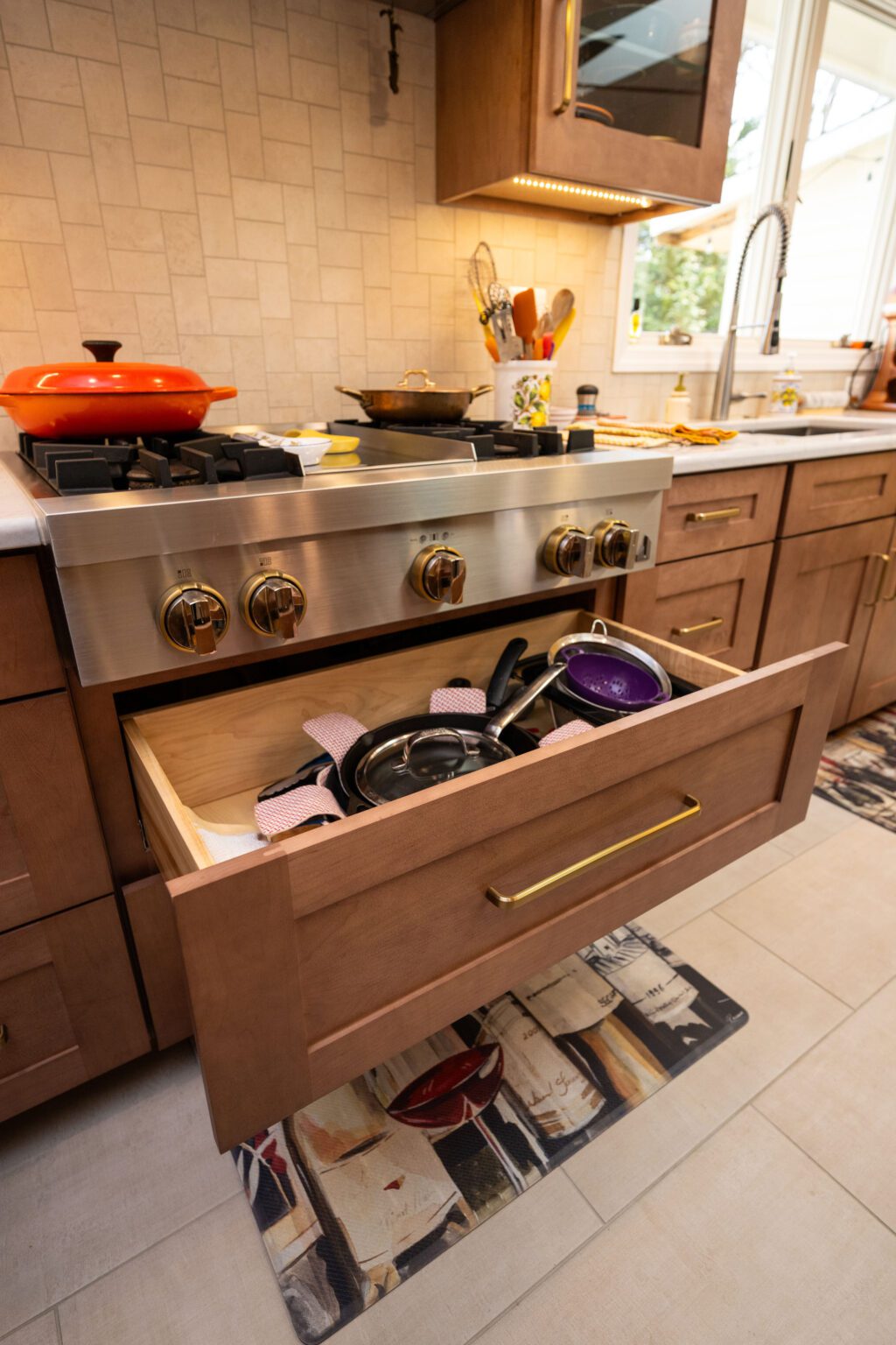 Image by Lesnick Photo A kitchen stove with an open drawer underneath, containing pots, pans, and lids. The countertop holds various kitchen items, and there is a window in the background.