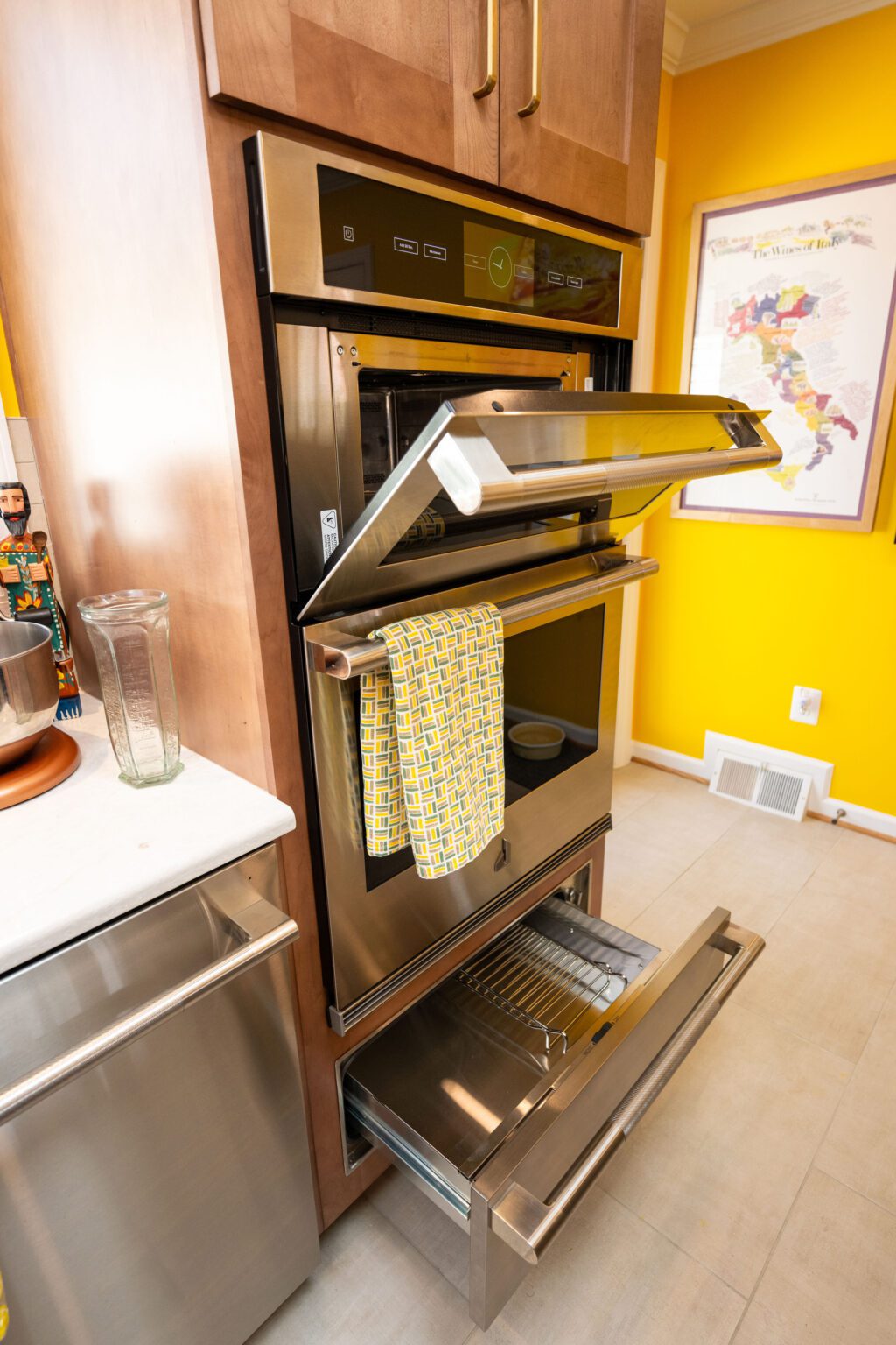 Image by Lesnick Photo Double wall oven with the top oven door open and a yellow-patterned towel hanging on the handle; lower warming drawer is also open in a yellow kitchen.