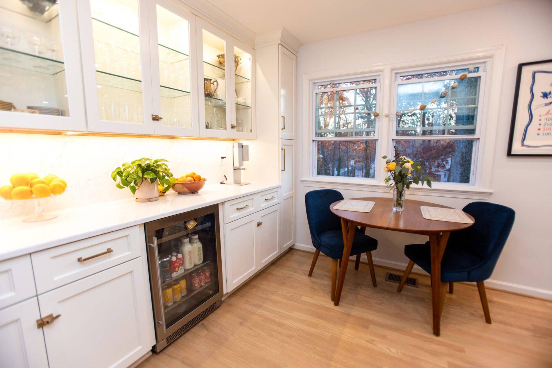 A bright kitchen with white cabinets, a glass-door mini fridge, fruit and a plant on the counter, plus a small wooden table and two blue chairs by the window—perfect inspiration if you’re wondering how much does a kitchen remodel cost in Northern Virginia.