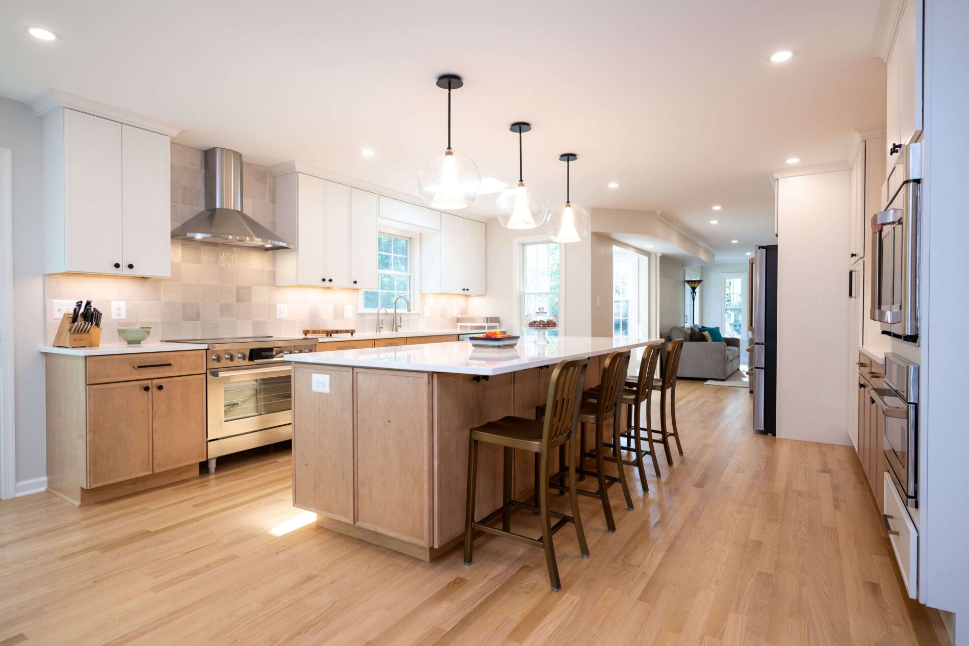 Modern kitchen with light wood floors, white cabinets, stainless steel appliances, a large island with bar stools, pendant lighting, and an open view to a living area—discover how much does a kitchen remodel cost in Northern Virginia.