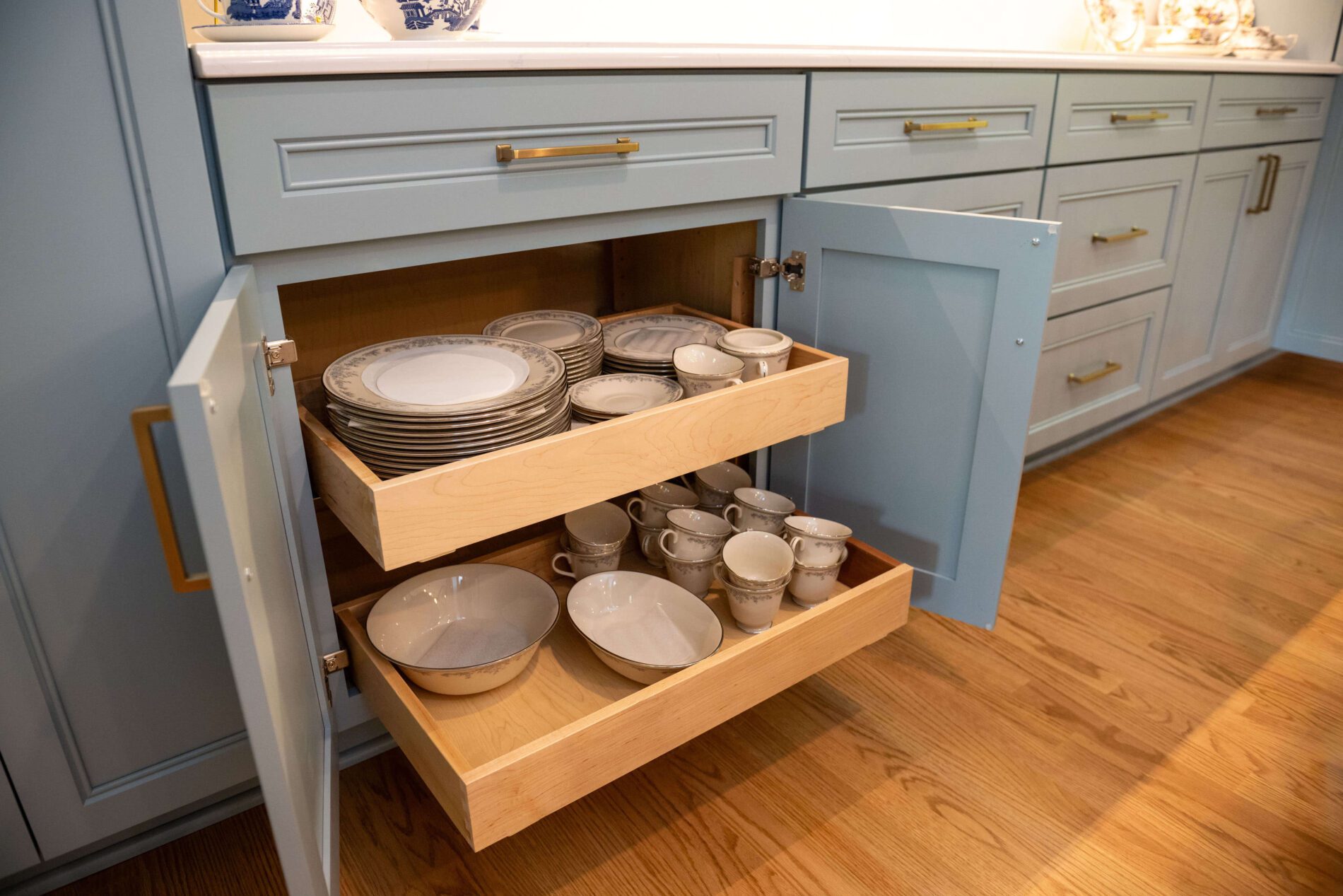 Two wooden pull-out drawers in a blue kitchen cabinet hold stacks of plates, bowls, and cups, neatly organized on a wooden floor—a smart storage idea if you’re wondering how much does a kitchen remodel cost in Northern Virginia.