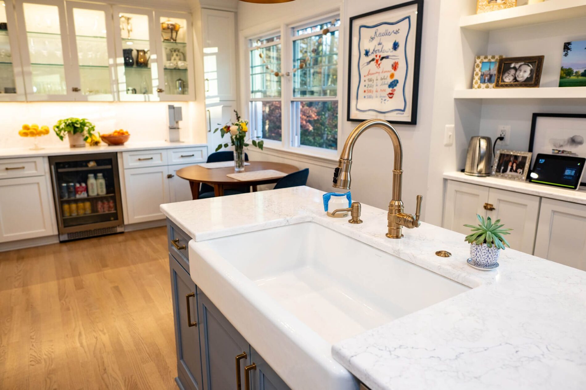 Modern kitchen with a white farmhouse sink, gold faucet, marble countertops, glass-front cabinets, a dining table, and natural light from large windows. Wondering how much does a kitchen remodel cost in Northern Virginia? This style offers inspiration.