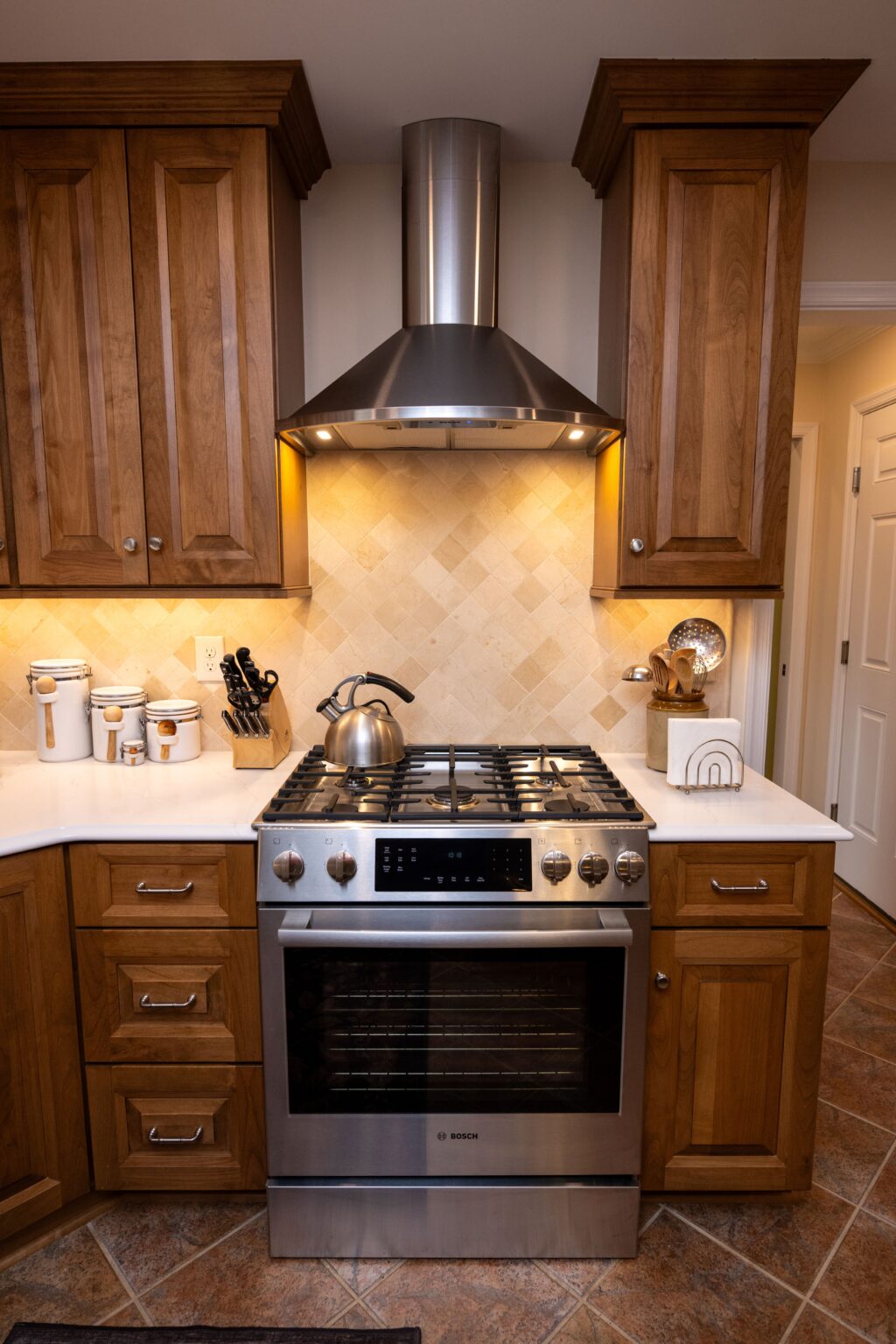 A modern kitchen features a stainless steel gas stove with a kettle, wooden cabinets, a range hood, and various kitchen utensils on the countertops.