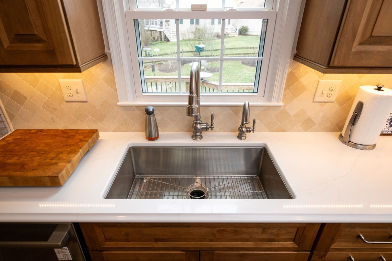 A modern kitchen sink with a metal faucet, soap dispenser, and paper towel holder on a white countertop with wooden cabinets and a window overlooking a backyard.